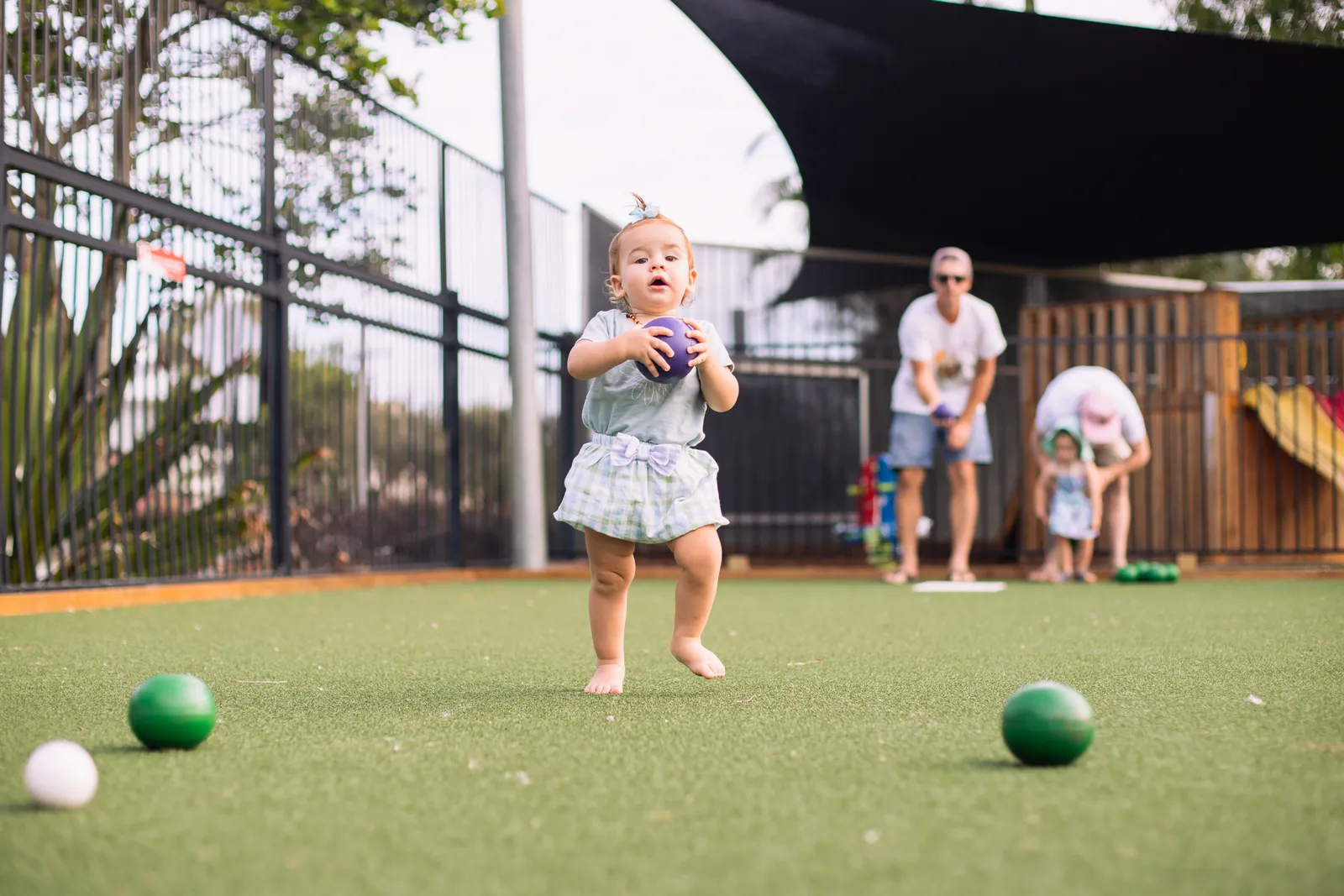 A toddler playing barefoot bowls with family on the green